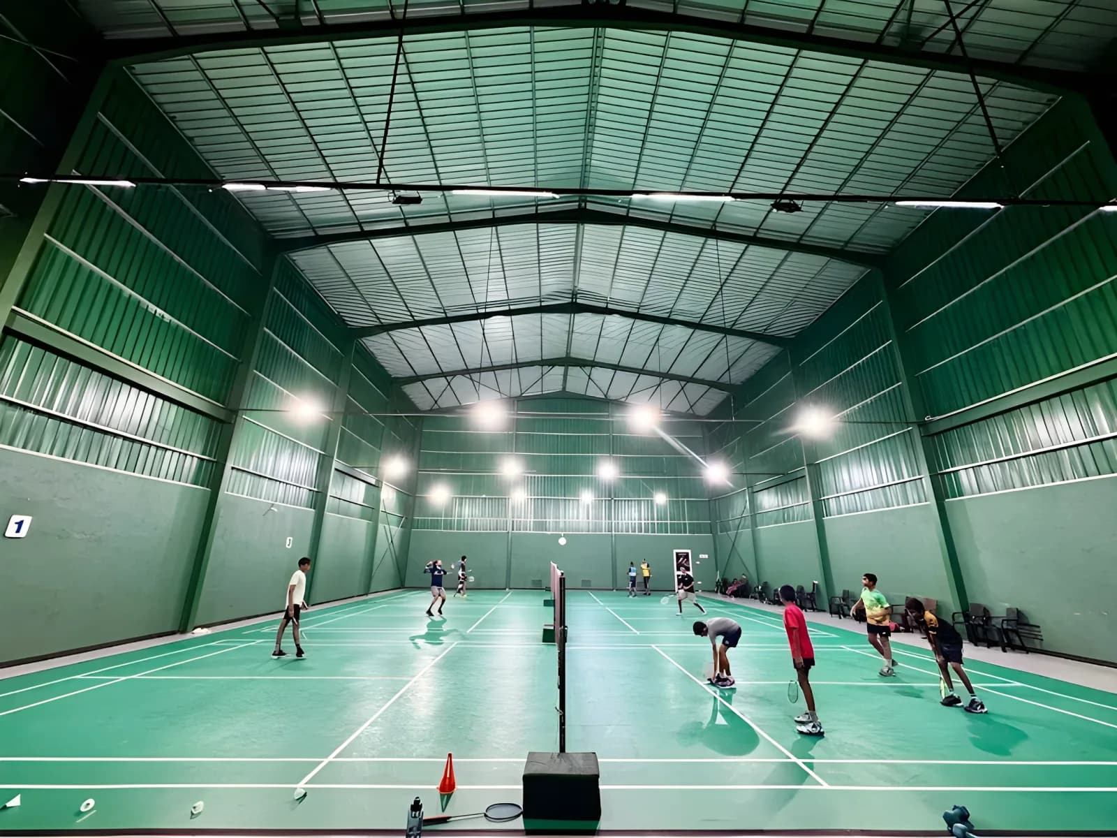 Players on three green synthetic courts at C & S Badminton Complex, Galle, under a high green steel-truss roof lit by rows of overhead lamps