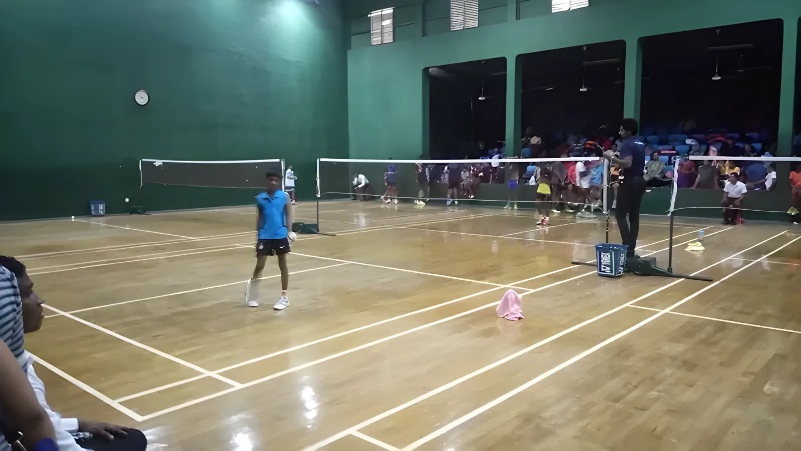 Juniors playing a tournament across several courts at Welagedara Stadium, Kurunegala, with green walls and spectators seated along the far side