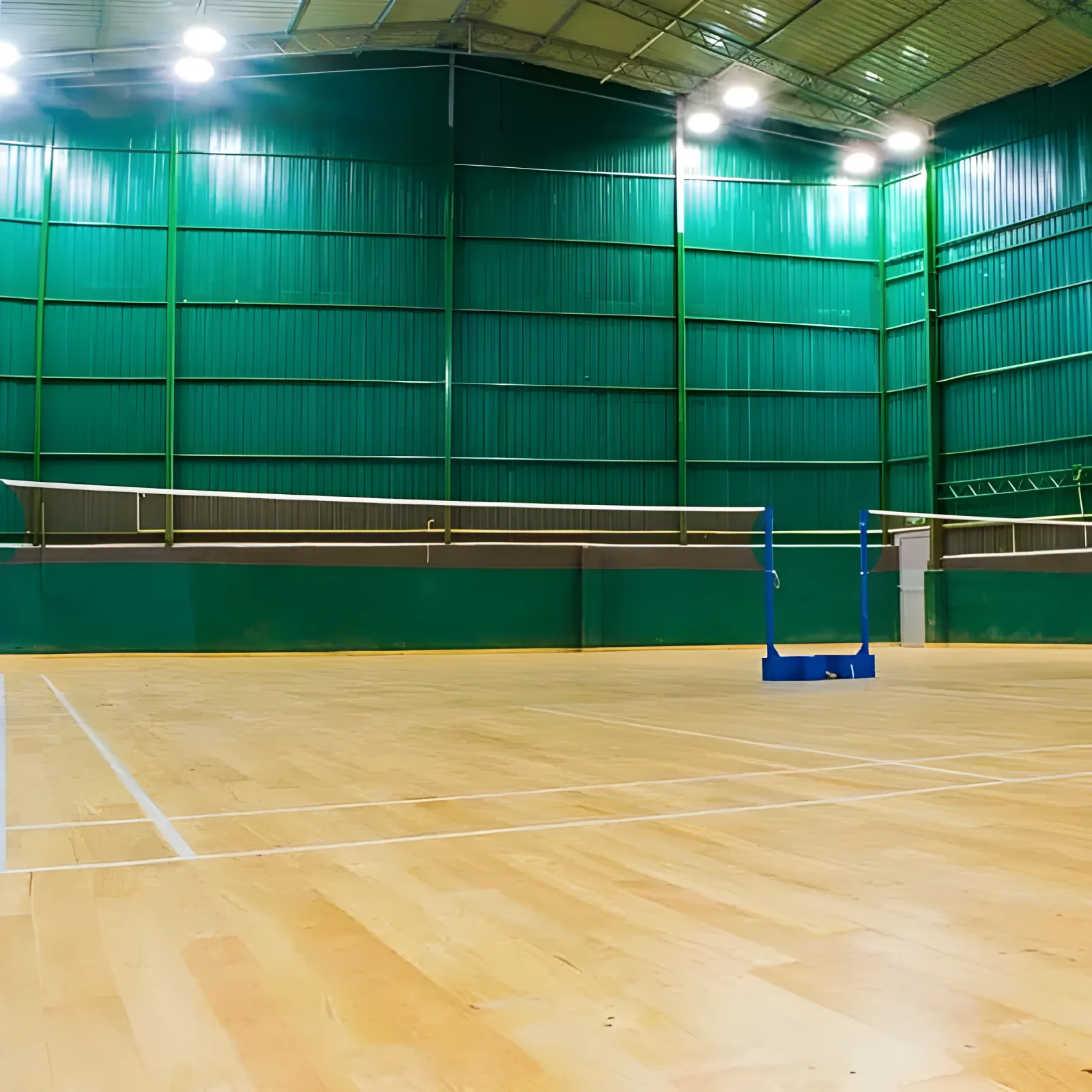 Empty wooden badminton court at Axion Sports Complex, Kandy, net up under a high green corrugated steel shed lit by overhead floodlights