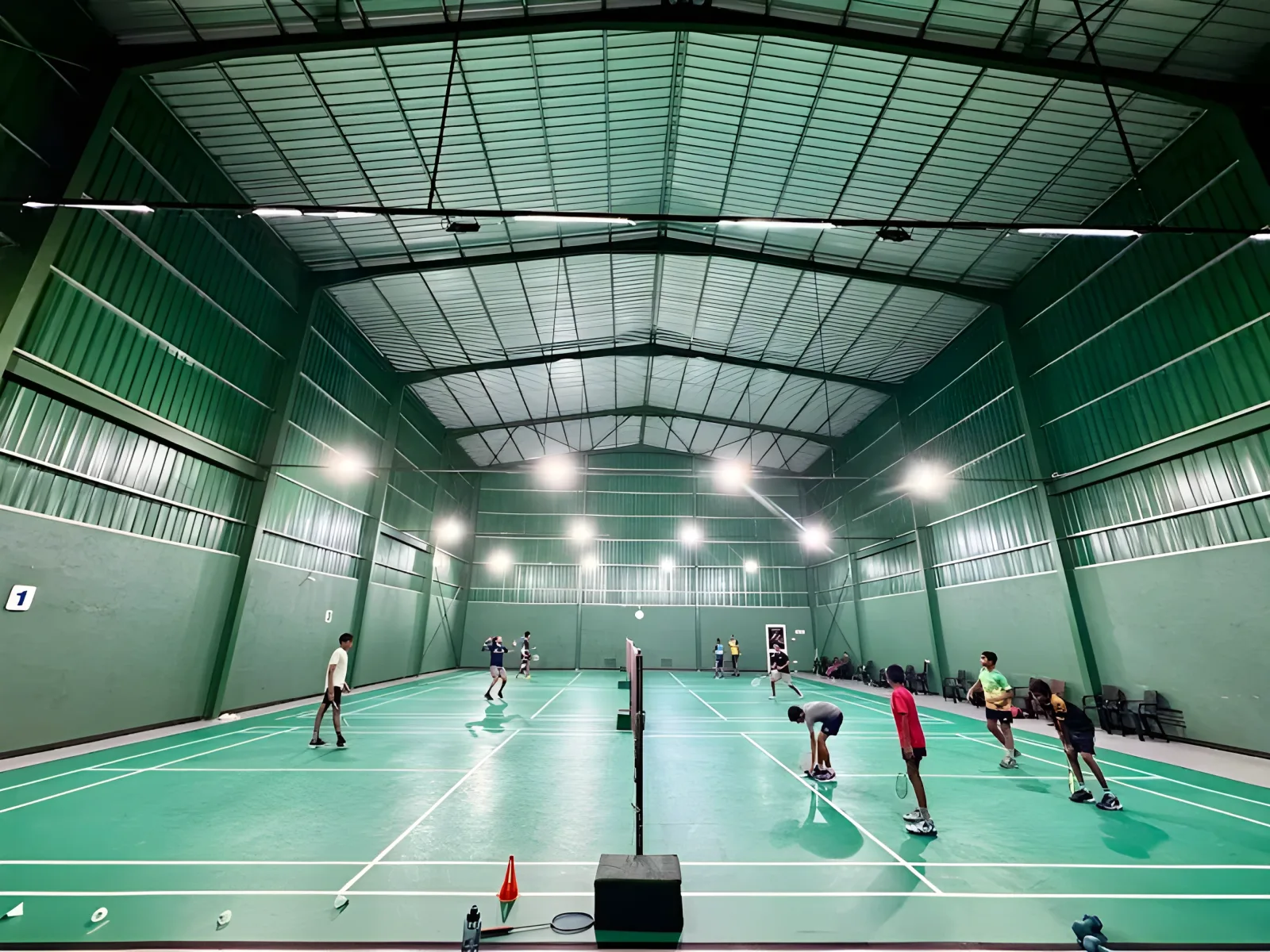 Players on three green synthetic courts at C & S Badminton Complex, Galle, under a high green steel-truss roof lit by rows of overhead lamps