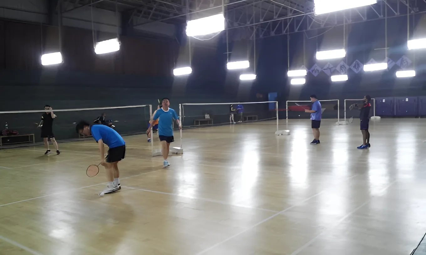 Players in casual doubles on pale wooden courts at D.S. Senanayake College Indoor Sports Complex, Colombo 07, several nets up under a high steel truss roof