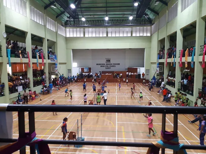 Junior tournament under way on the wooden floor at Kandy Municipal Indoor Stadium, several courts in play at once, seen from a packed first-floor spectator gallery