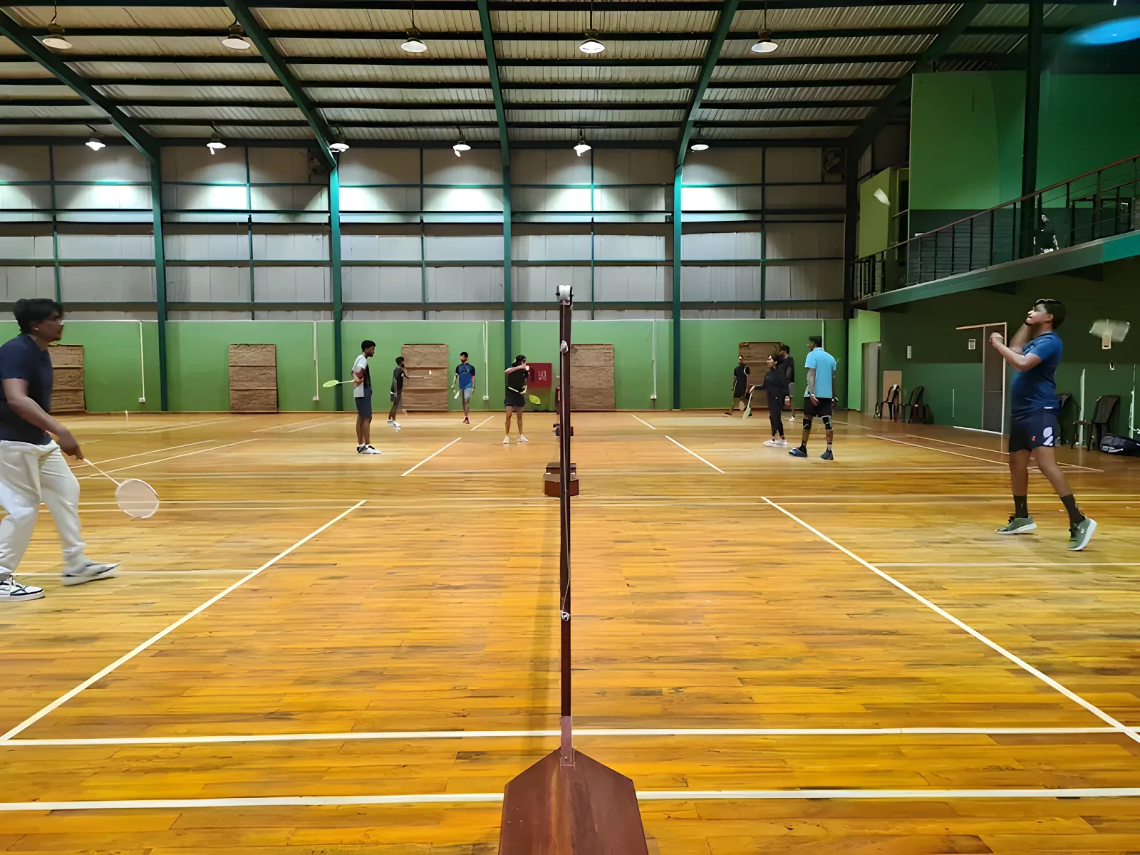 Players on several wooden courts at Makro Sports Arena, Gampaha, in a tall green-walled hall with a viewing balcony along one side