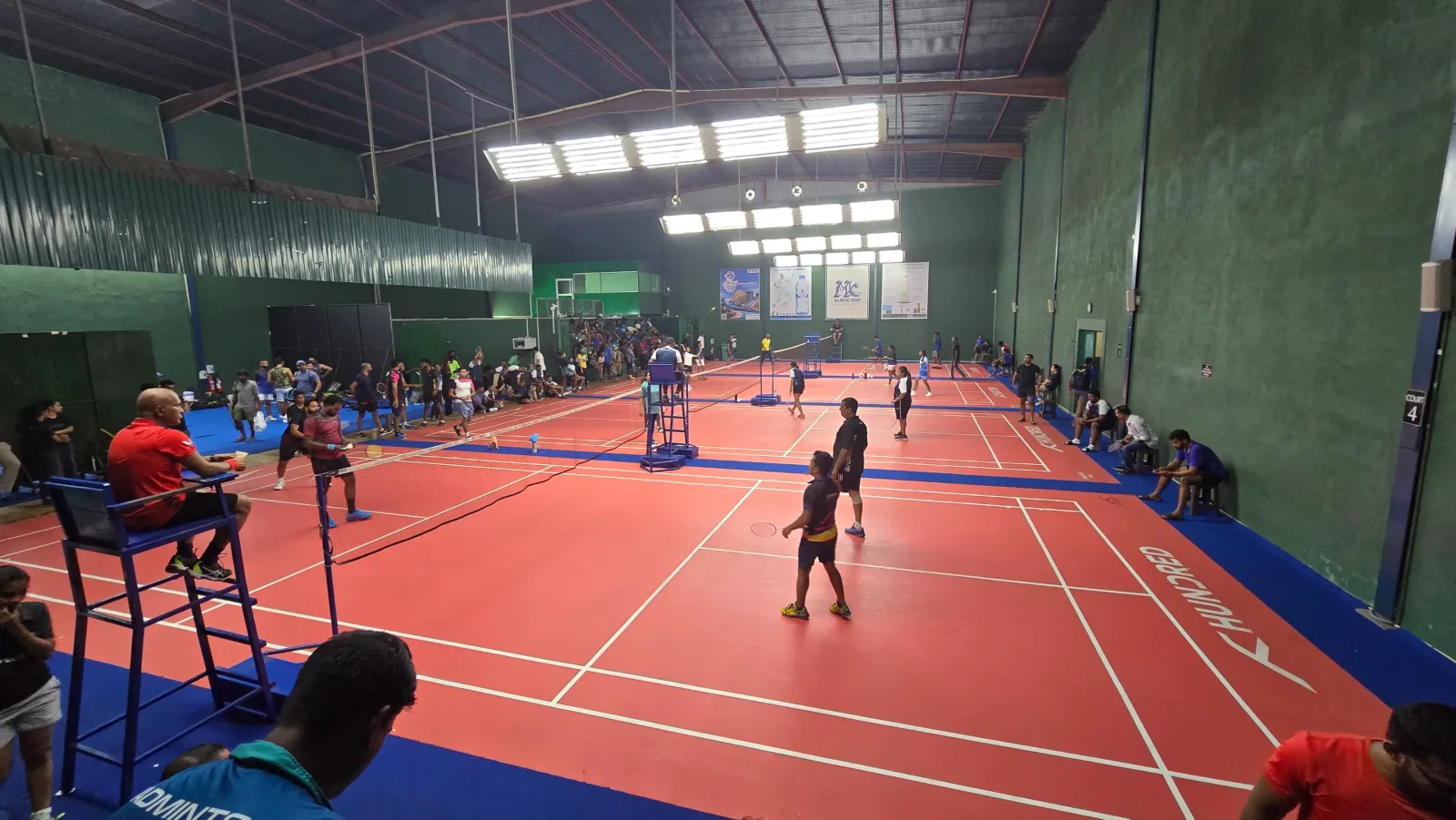 Players on red and blue synthetic courts at MBA Badminton Courts, Colombo 7, with umpire chairs and spectators along the green walls during a tournament