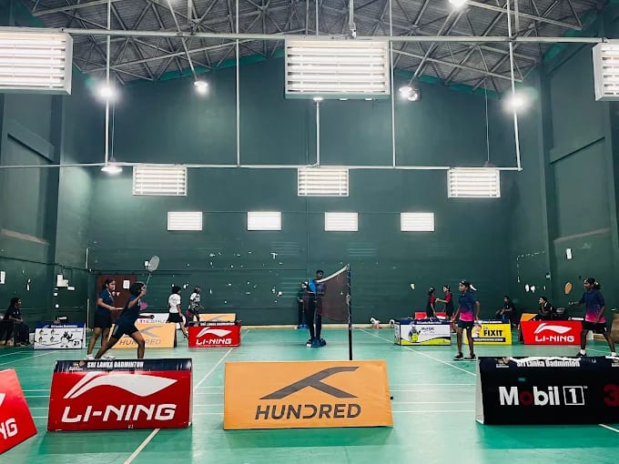 Players in a doubles rally on green synthetic carpet courts at Sri Lanka Badminton, Colombo 07, with sponsor boards along the side lines under a high steel truss roof