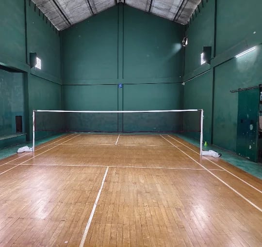 Single wooden badminton court at Super Sport, Nawala, net up between dark green walls under a pitched roof with exposed timber trusses and wall-mounted floodlights
