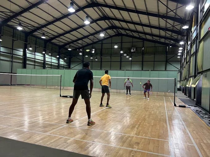 Players mid-rally on the sprung wooden courts at Zimantra Leisure Centre, Battaramulla, under a curved steel roof with green side panelling